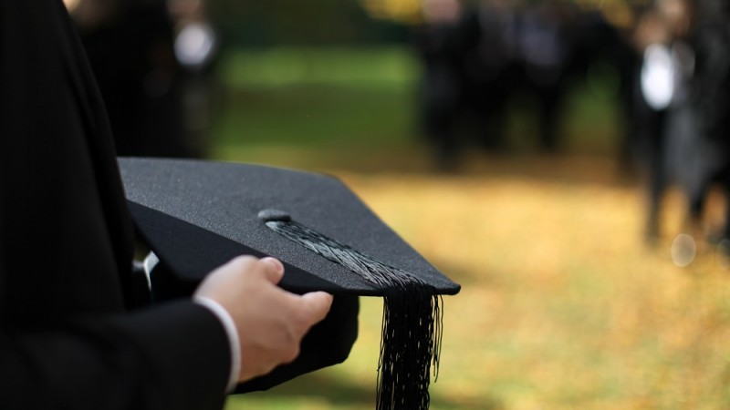 &Agrave;esquerda, bra&ccedil;o e m&atilde;o direita de uma pessoa vestida com toga segura um capelo. Ao fundo, com desfoque, grama e outras pessoas vestidas com toga.