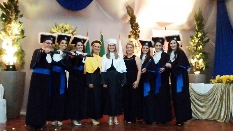 Formandos vestindo toga. Ao centro, a vice-reitora, a paraninfa e a funcion&aacute;ria homenageada.