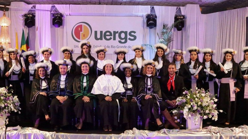 Sobre o palco e em p&eacute;, turma de formandos vestindo toga; em frente a eles e sentados, a reitora, paraninfos e homenageados. Ao fundo, painel com o logotipo da Uergs.
