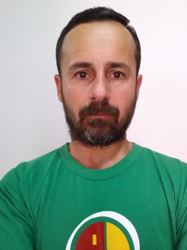 Homem de pele clara, cabelo curto preto, barba e bigode grisalhos. Tem semblante sério e veste camiseta verde com o logo da Uergs.