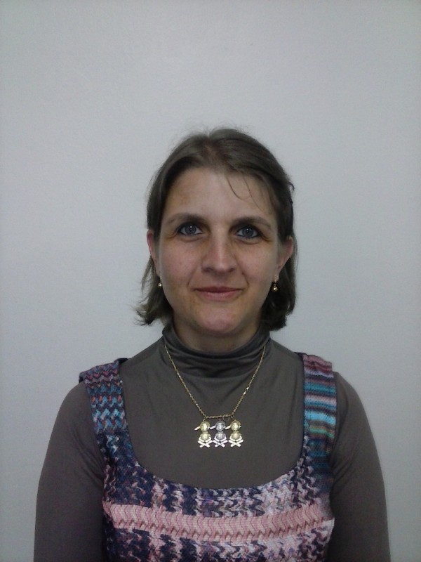 Mulher de pele clara, olhos azuis e cabelos louros, lisos, em comprimento chanel. Veste blusa cinza de gola alta, sobreposta com regata estampada e usa colar durado com pingente de três crianças.