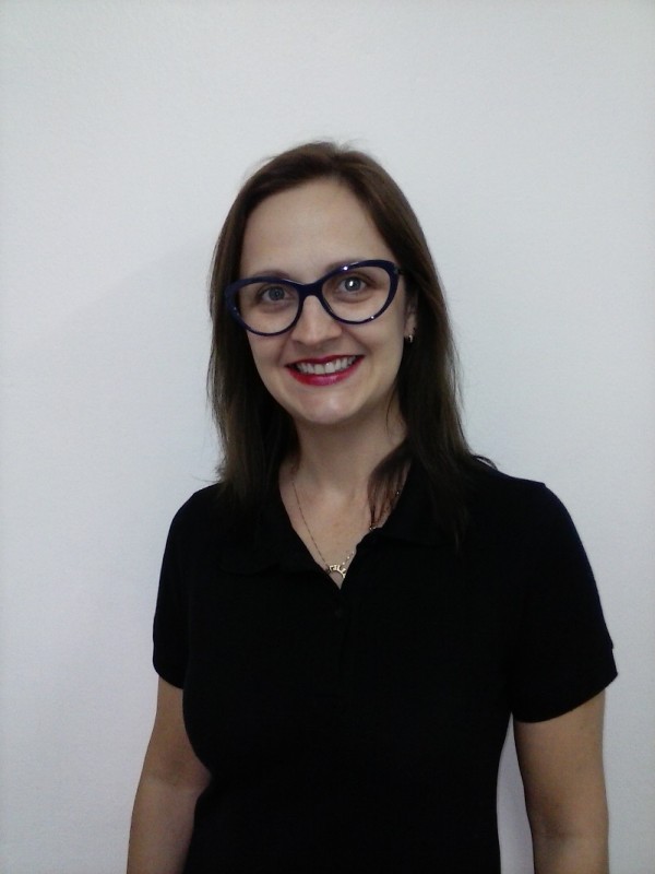 Mulher sorridente de pele clara, cabelos castanhos, lisos e compridos. Veste blusa preta e usa óculos de graus pretos com batom vermelho.