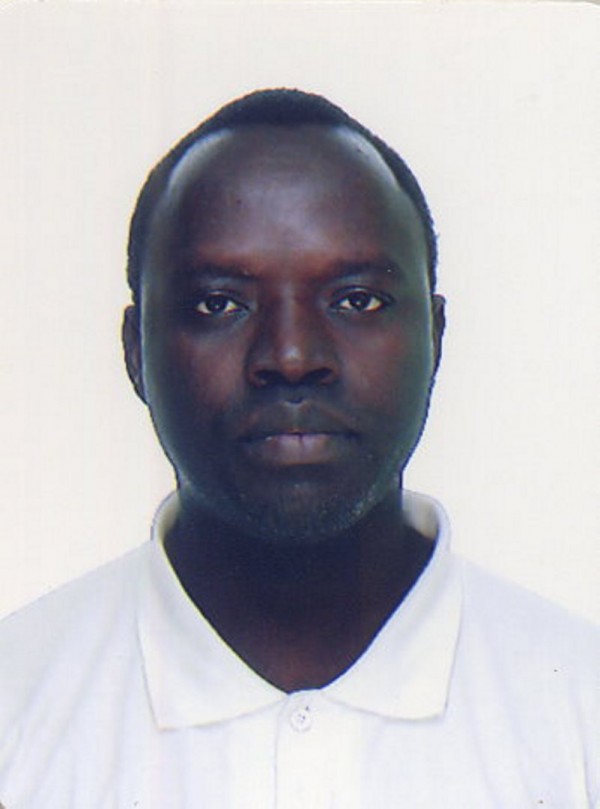 Homem de pele negra, cabelo curto preto e semblante sério. Veste camisa polo branca.