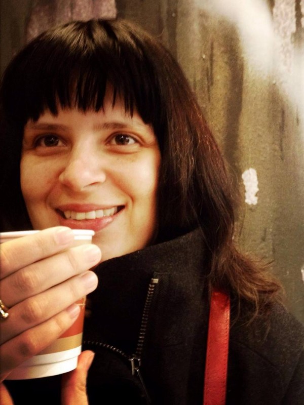 Mulher de pele clara, cabelos escuros médios, com franja, olhos escuros. Sorri enquanto segura um copo de café.