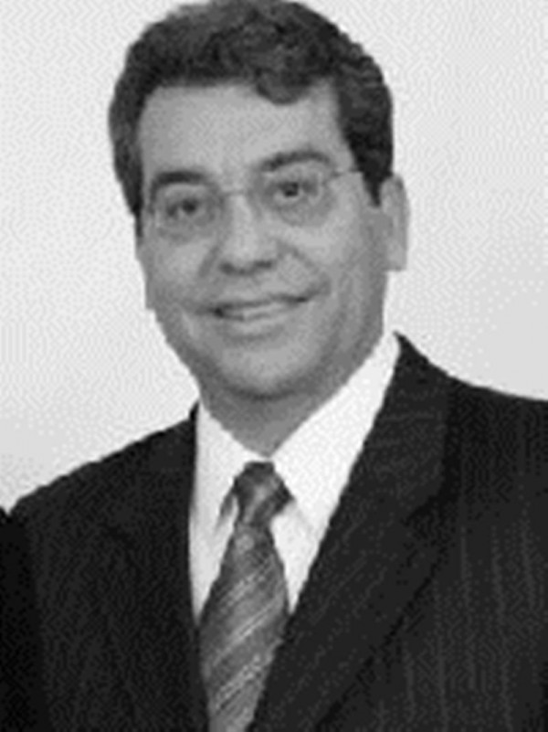 Foto em preto e branco de homem sorridente de cabelos escuros curtos e pele clara. Usa óculos de grau, terno e gravata.