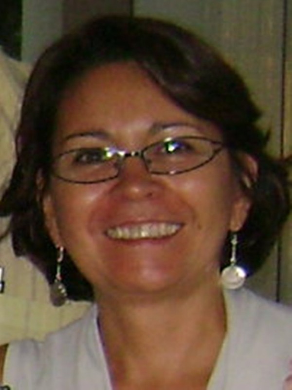 Mulher de pele morena clara, cabelo chanel escuro, olhos escuros. Usa óculos de grau e está sorridente.