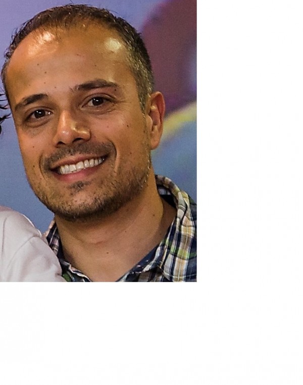 Homem sorridente de pele morena, cabelos pretos, barba e bigode pouco volumosos.