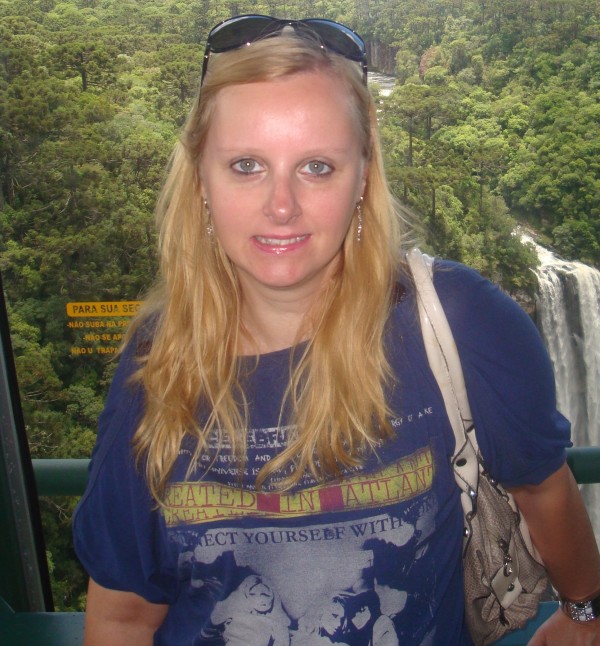 Mulher sorridente de pele clara, olhos azuis, cabelos louros, lisos e compridos. Usa óculos de sol prendendo os cabelos.