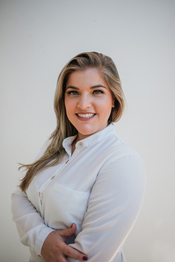 Mulher sorridente de pele e olhos claros, cabelos louros, lisos e compridos. Está de perfil com os braços cruzados e veste camisa branca.