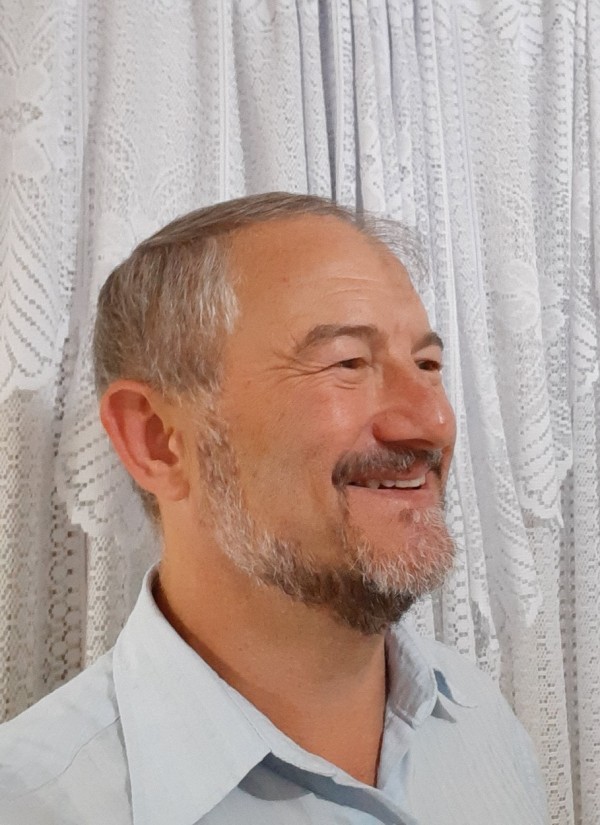 Homem sorridente de pele clara, barba, bigode e cabelo grisalhos. Está de perfil e veste camisa azul clara.