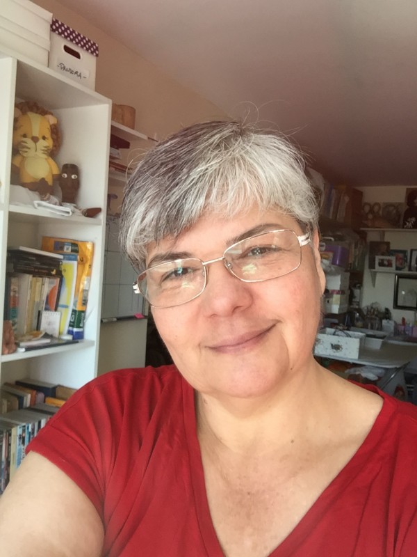 Mulher de pele clara, cabelos grisalhos e curtos. Usa óculos de grau, veste blusa vermelha e sorri ligeiramente.