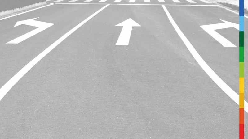 Imagem em preto e branco de uma rua asfaltada de tr&ecirc;s vias, com setas brancas pintadas no ch&atilde;o, sendo uma para a esquerda, uma para a frente e uma para a direita. Na lateral direita da imagem, uma faixa colorida vertical.