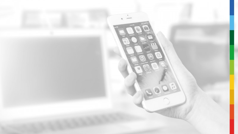 Imagem em preto e branco de uma m&atilde;o segurando um smartphone, com um notebook ao fundo. Na lateral direita da imagem, uma faixa colorida vertical.