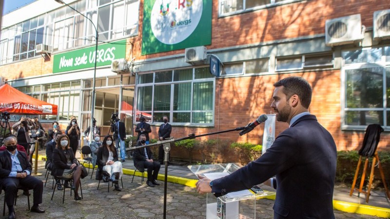 Foto colorida. Em primeiro plano, &agrave; direita, o governador Eduardo Leite, com terno escuro, faz seu discurso ao microfone, diante de um p&uacute;lpito acr&iacute;lico. Em segundo plano, pessoas convidadas est&atilde;o sentadas &agrave; sua frente. Ao fundo, pr&eacute;dio de tijolos &agrave; vista e grandes janelas, que &eacute; a nova sede da Reitoria da Uergs. 