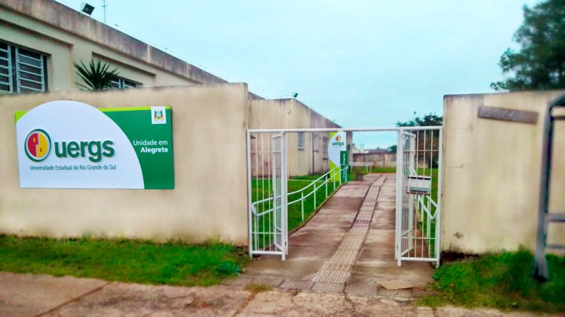 Em primeiro plano, foto de um muro claro e um port&atilde;o, com a placa de identifica&ccedil;&atilde;o da Uergs &agrave; esquerda. Ao fundo, pr&eacute;dio de um andar na mesma cor do muro e totem da Uergs.