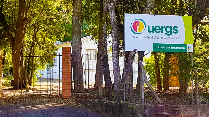 Em primeiro plano, a placa de identifica&ccedil;&atilde;o da Uergs e algumas &aacute;rvores. Ao fundo, pr&eacute;dio branco de um andar.