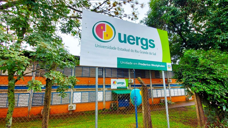 &Agrave; frente, em primeiro plano, placa de identifica&ccedil;&atilde;o da Uergs. Ao fundo, pr&eacute;dio de 2 andares com paredes de tijolos &agrave; vista e detalhes em azul.