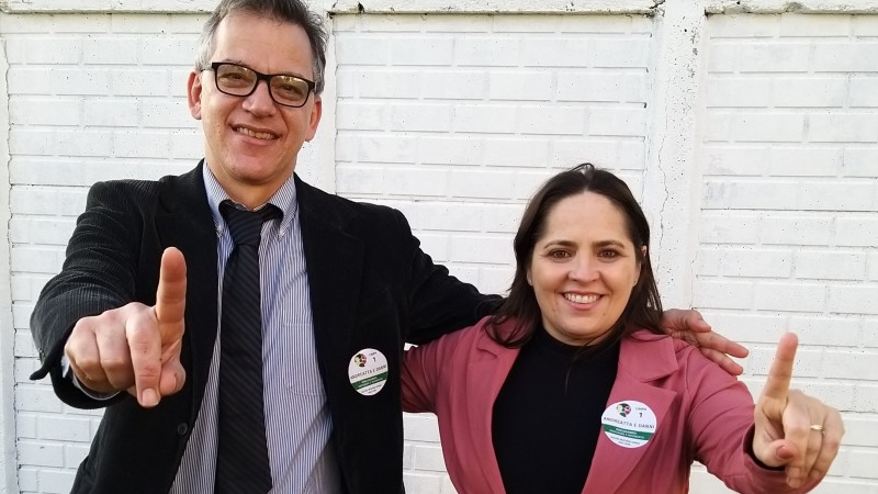 Foto colorida de um homem e uma mulher posando sorridentes e fazendo o sinal de 1 com o dedo indicador. Ele tem a pele clara, cabelo grisalho, usa &oacute;culos de grau e veste terno e gravata. Ela &eacute; morena clara, cabelos lisos e pretos, veste blusa preta com terninho rosa.