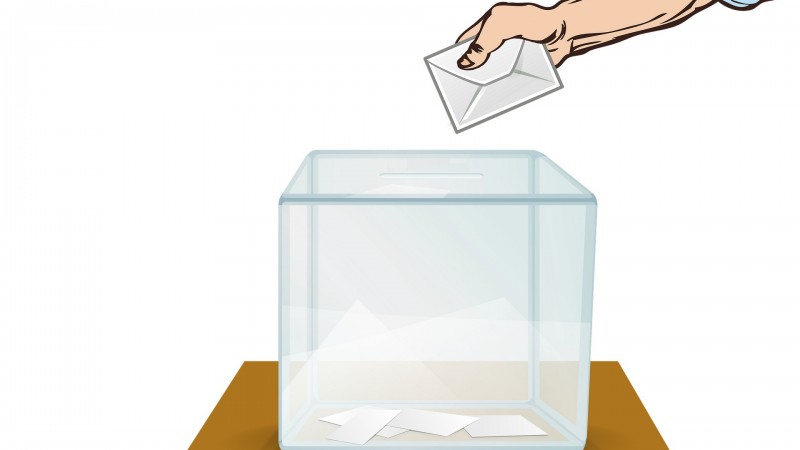 Imagem com fundo branco. Ao centro, ilustra&ccedil;&atilde;o de uma urna de vidro transparente sobre uma mesa de madeira, com peda&ccedil;os de papel dentro. Sobre ela, a m&atilde;o de um homem segurando um envelope.