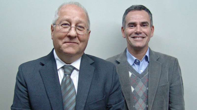 Dois homens de pele clara e sorridentes, vestindo ternos de cores neutras. O da esquerda tem cabelos brancos, ligeiramente calvo e usa &oacute;culos de grau. O da direita tem cabelos curtos e grisalhos.