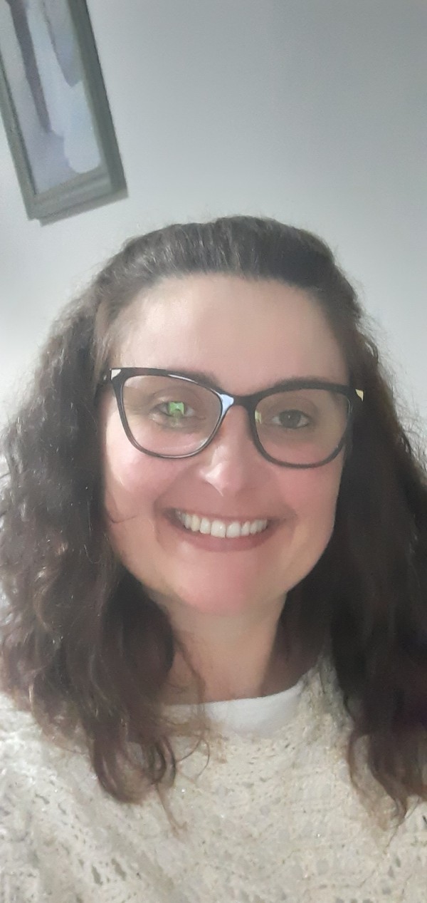 Mulher sorridente de pele morena clara com óculos de grau, olhos castanhos e cabelos castanhos ondulados.