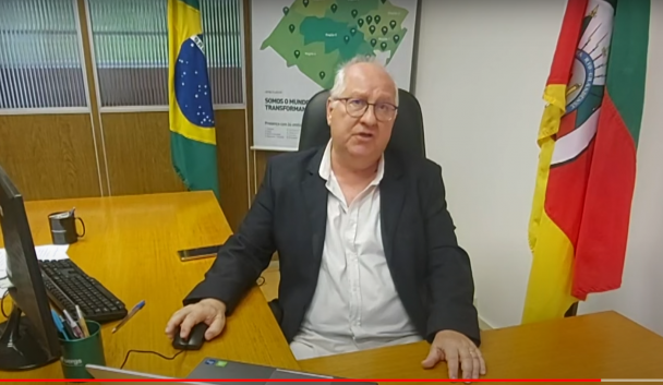 Homem de pele clara, cabelos brancos curtos, sentado em seu Gabinete. Veste camisa branca e terno preto. Atrás dele e em lados opostos, a bandeira do Brasil e do RS.