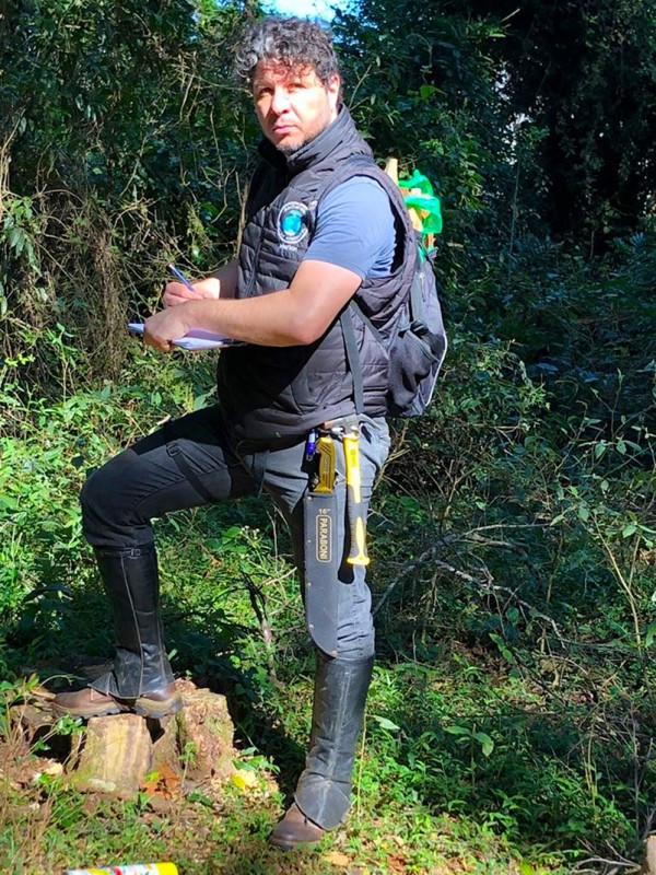 Homem alto com cabelos pretos curto, barba, de pele clara. Veste roupas adequadas ao trabalho em campo e posa em meio à vegetação segurando uma prancheta e uma caneta.