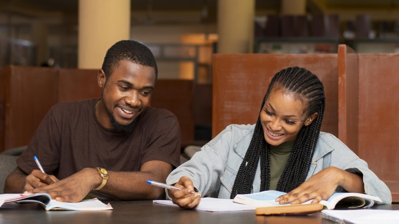 Dois jovens negros sorridentes (um homem e uma mulher), sentados olhando para livros e cadernos, com canetas nas m&atilde;o. Ele tem cabelos curtos, bigode e barba, e veste camiseta marrom. Ela tem cabelos compridos com tran&ccedil;as nag&ocirc; e veste jaqueta jeans.